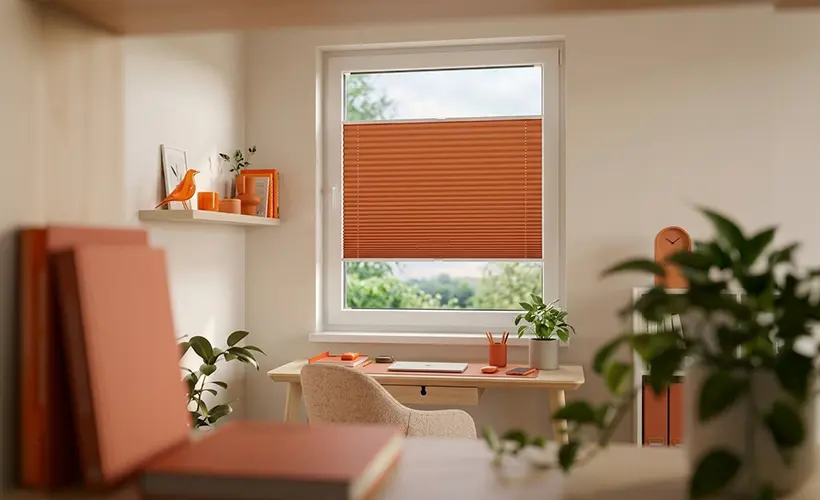 A home office with an orange pleated blind on the window and a bright arrangement in natural shades
